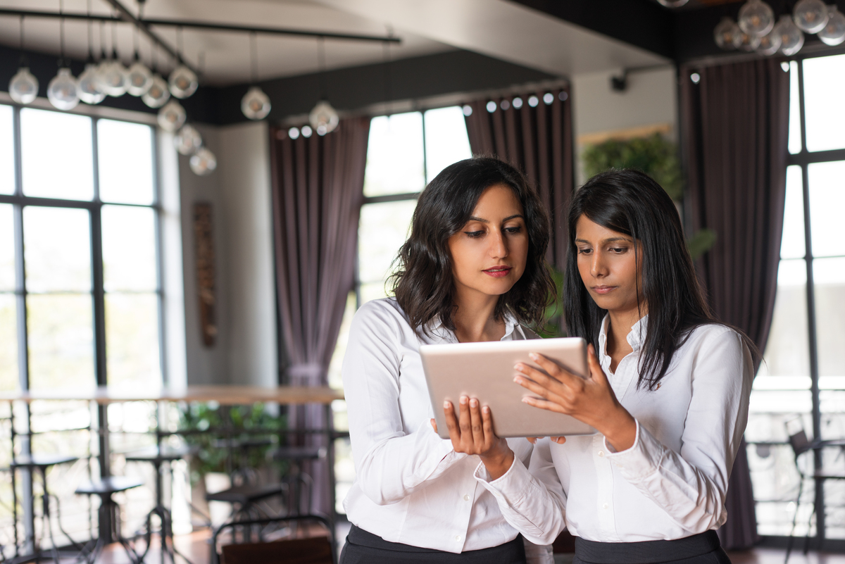 Two focused female colleagues using tablet computer in cafe. They are standing with cafe interior and windows in background. Technology concept. Front view.